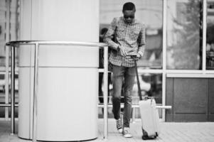 homem afro-americano de camisa quadriculada, com mala e mochila. viajante negro contra a rodoviária. foto