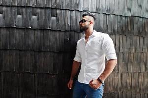 elegante modelo de homem árabe alto na camisa branca, jeans e óculos de sol posou contra a parede de madeira interior. barba cara árabe atraente. foto