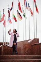 jovem elegante linda mulher afro-americana na rua, vestindo casaco de roupa de moda, contra bandeiras de diferentes países do mundo, com telefone celular na mão. foto