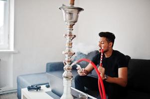 homem indiano bonito e elegante em preto sentado na sala, fumando cachimbo de água, olhando no telefone com o laptop. foto