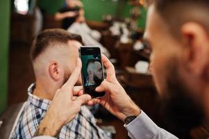 homem barbudo bonito na barbearia, barbeiro no trabalho, fazendo foto em seu telefone.