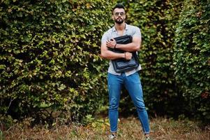 homem de barba árabe alto na moda usar camisa, jeans e óculos de sol posou no parque e segure a mala. foto