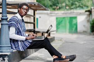 homem africano em roupas tradicionais e óculos com laptop trabalhando ao ar livre. foto