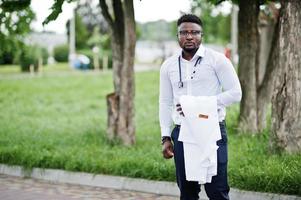 jovem médico americano africano segura jaleco branco por lado com um estetoscópio posado ao ar livre. foto