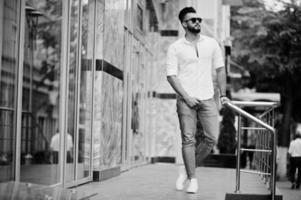 elegante modelo de homem árabe alto na camisa branca, jeans e óculos de sol posou na rua da cidade. barba cara árabe atraente. foto