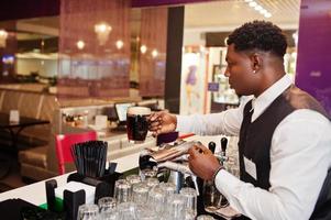 barman americano africano no bar segura cerveja escura fresca no vidro no pub. foto