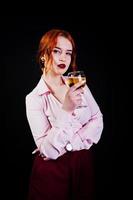 linda garota ruiva de blusa rosa e saia vermelha com copo de vinho na mão isolada em preto. foto