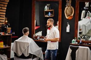 jovem barbudo cortando o cabelo pelo cabeleireiro enquanto está sentado na cadeira na barbearia. alma de barbeiro. foto