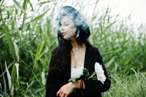 garota fumante sensual toda em chapéu e lábios pretos, vermelhos. mulher dramática gótica segura flor de crisântemo branco e fumando em junco comum. foto