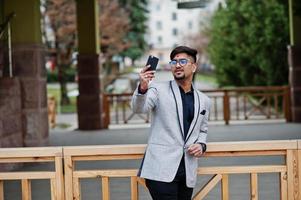 homem de negócios indiano elegante com bindi na testa e óculos, use terno cinza posou ao ar livre e fazendo selfie no celular. foto