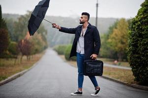 homem de barba árabe alto na moda usa casaco preto com guarda-chuva e bolsa posada no dia de chuva. foto