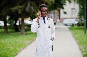 elegante médico americano africano com estetoscópio e jaleco, em óculos posados ao ar livre. foto