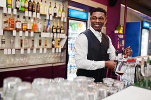 barman americano africano no bar derramando da torneira de cerveja fresca no copo no pub. foto