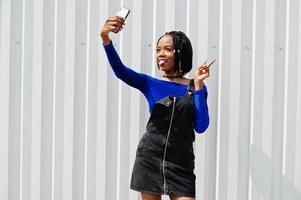 mulher africana de saia de macacão jeans e vestido azul posou contra a parede de aço branca e fazendo selfie no telefone. foto