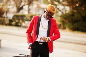 modelo de homem afro-americano de moda no terno vermelho, com destaques de cabelo e bolsa posou na rua e olhando para seus relógios. foto