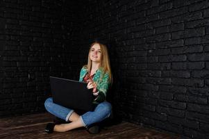 menina loira elegante jaqueta e jeans com laptop contra parede de tijolo preto no estúdio. foto