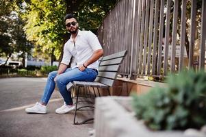 elegante modelo de homem árabe alto na camisa branca, jeans e óculos de sol posou na rua da cidade. barba cara árabe atraente sentado no banco. foto