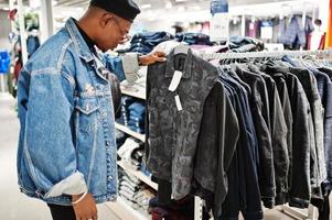 homem afro-americano casual elegante na jaqueta jeans e boina preta na loja de roupas olhando na nova camisa militar. foto