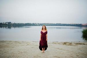 mulher loira sensual descalça no vestido vermelho marsala posando contra o lago na areia. foto