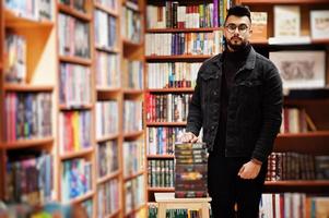 homem alto estudante árabe inteligente, use jaqueta jeans preta e óculos, na biblioteca com pilha de livros. foto