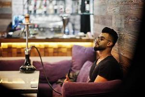 homem árabe de barba elegante de óculos e camiseta preta fumando narguilé bar interno. modelo árabe descansando. foto