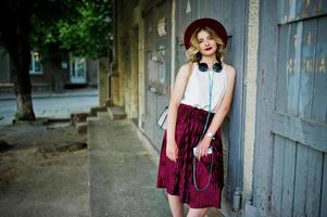 menina modelo loira elegante e bonita na saia de veludo vermelho elegante, blusa branca e chapéu, posou com telefone e fones de ouvido. foto