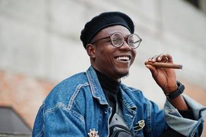 feche o retrato do homem afro-americano na jaqueta jeans, boina e óculos, fumando charuto. foto