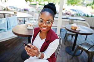 garota afro-americana usa óculos com celular sentado no caffe ao ar livre. foto