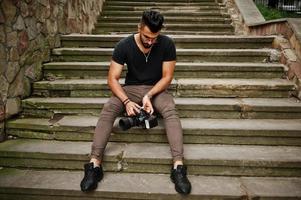 Fotógrafo de homem macho de barba árabe linda e bonita em óculos e camiseta preta com câmera profissional nas mãos. foto