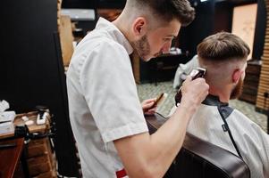 jovem barbudo cortando o cabelo pelo cabeleireiro enquanto está sentado na cadeira na barbearia. alma de barbeiro. foto