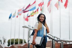 mulher estudante africana posou com mochila e itens escolares no pátio da universidade, contra bandeiras de diferentes países. foto