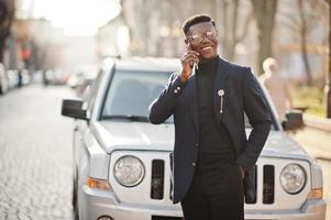 homem afro-americano incrivelmente bonito usa blazer azul com broche, gola alta preta e óculos posados na rua. cara negro na moda falando no telefone contra o carro. foto