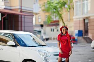 linda e magra garota afro-americana de vestido vermelho com dreadlocks posou ao ar livre no carro branco pequeno de fundo de rua. elegante modelo preto. foto