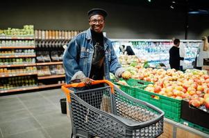 homem afro-americano casual elegante na jaqueta jeans e boina preta na seção orgânica do supermercado com carrinho de compras. foto