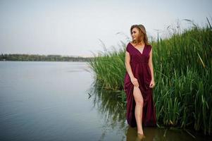 loira sensual em pé de vestido vermelho marsala na água do lago com juncos. foto