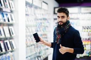 comprador de cliente homem de barba indiana na loja de telefonia móvel escolhe seu novo smartphone e mostra o polegar para cima. conceito de povos e tecnologias do sul da Ásia. loja de celulares. foto