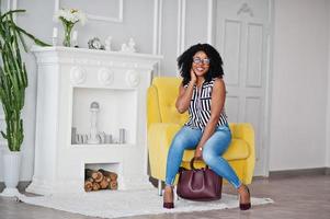 linda mulher afro-americana com cabelo afro encaracolado e óculos, bolsa posada no quarto, sentado na cadeira amarela. foto