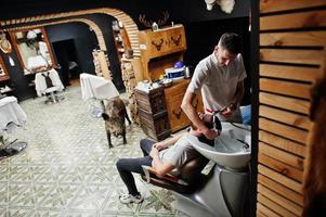 jovem barbudo lavando a cabeça pelo cabeleireiro enquanto está sentado na cadeira na barbearia. alma de barbeiro. foto