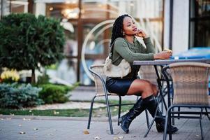 mulheres afro-americanas elegantes na moda na camisola verde e saia preta posou café ao ar livre, sentado à mesa com uma xícara de café. foto