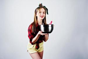 jovem dona de casa engraçada na camisa quadriculada e short amarelo pin up estilo com panela e colher de cozinha isolado no fundo branco. foto