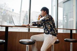 jovem freelancer indiano masculino sentado computador portátil aberto no café de fast food, bonito homem asiático falando telefone durante o trabalho no net-book no café confortável. foto