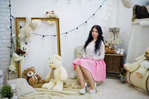 jovem morena de saia rosa e blusa branca posou interior contra quarto com urso de brinquedos. foto