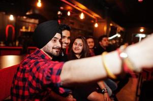 grupo de amigos indianos sentados no lounge bar, se divertindo e descansando, fumando narguilé e olhando em telefones celulares para fazer selfie. foto