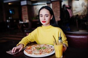 menina morena engraçada de suéter amarelo comendo pizza no restaurante. foto