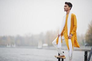 homem elegante indiano em roupas tradicionais amarelas com lenço branco posou ao ar livre contra o cais nebuloso do mar. foto