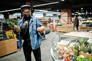 homem afro-americano casual elegante na jaqueta jeans e boina preta segurando cocos e abacaxi na seção orgânica de frutas do supermercado. foto