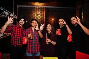 grupo de amigos indianos se divertindo e descansando na boate, bebendo coquetéis e mostrando os dedos ok juntos. foto