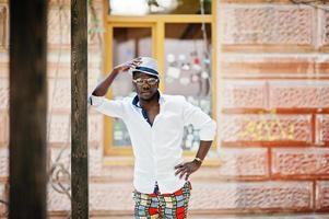 homem afro-americano elegante na camisa branca e calça colorida com chapéu e óculos posou ao ar livre. menino modelo preto elegante. foto