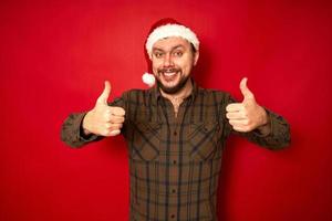 homem sorridente com chapéu de natal, roupas casuais mostra os polegares com as duas mãos isoladas no fundo vermelho do estúdio com espaço para texto. conceito - feriados, ano novo, promoção, descontos, oferta favorável foto