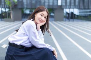 linda estudante asiática do ensino médio no uniforme escolar com aparelho nos dentes senta e sorri com confiança enquanto ela olha para a câmera alegremente com o prédio ao fundo. foto